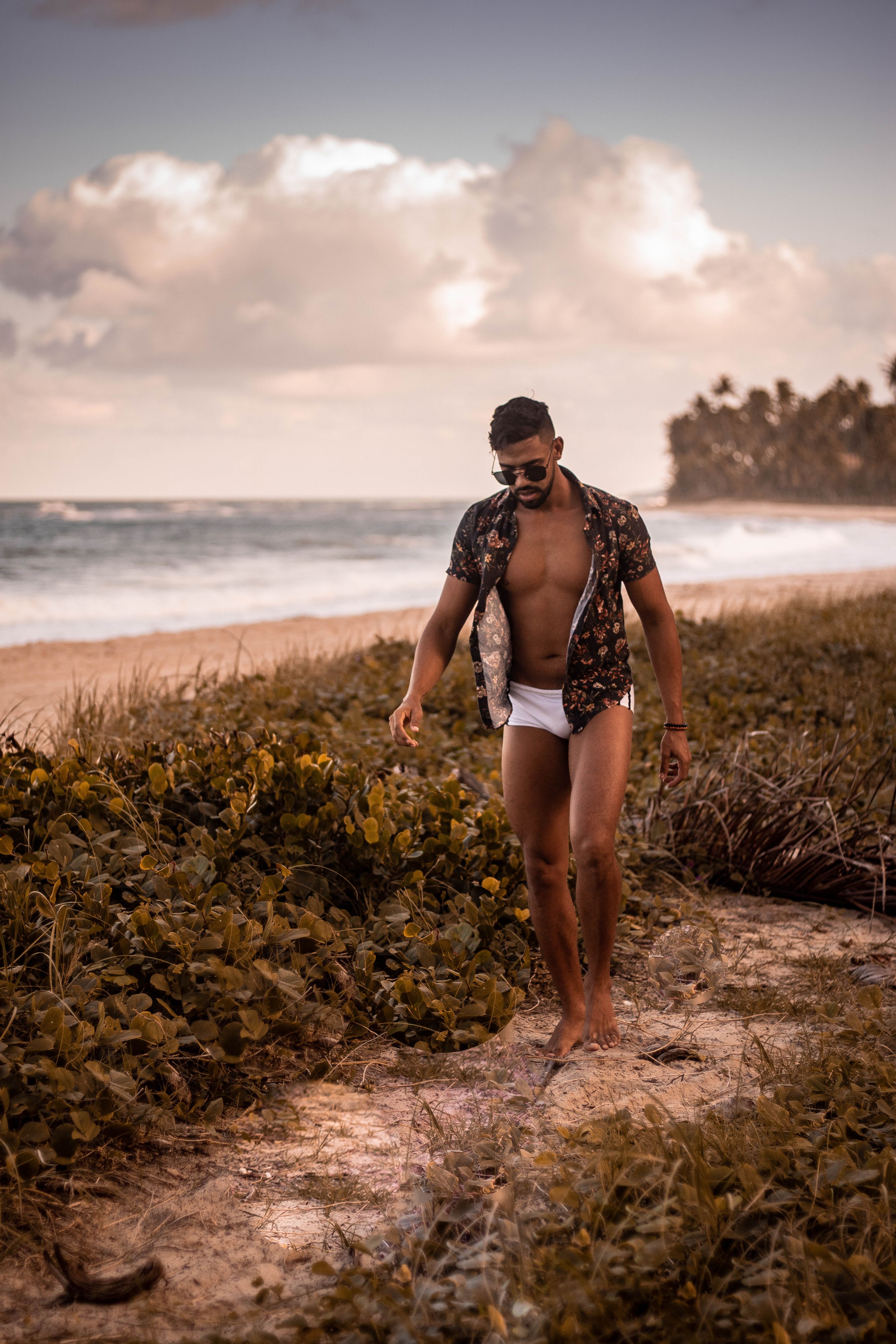 Um jovem modelo masculino caminha por uma praia cênica durante o pôr do sol, incorporando moda e estilo de verão.