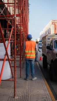 A construction worker in an orange safety vest stands by scaffolding on a city street.