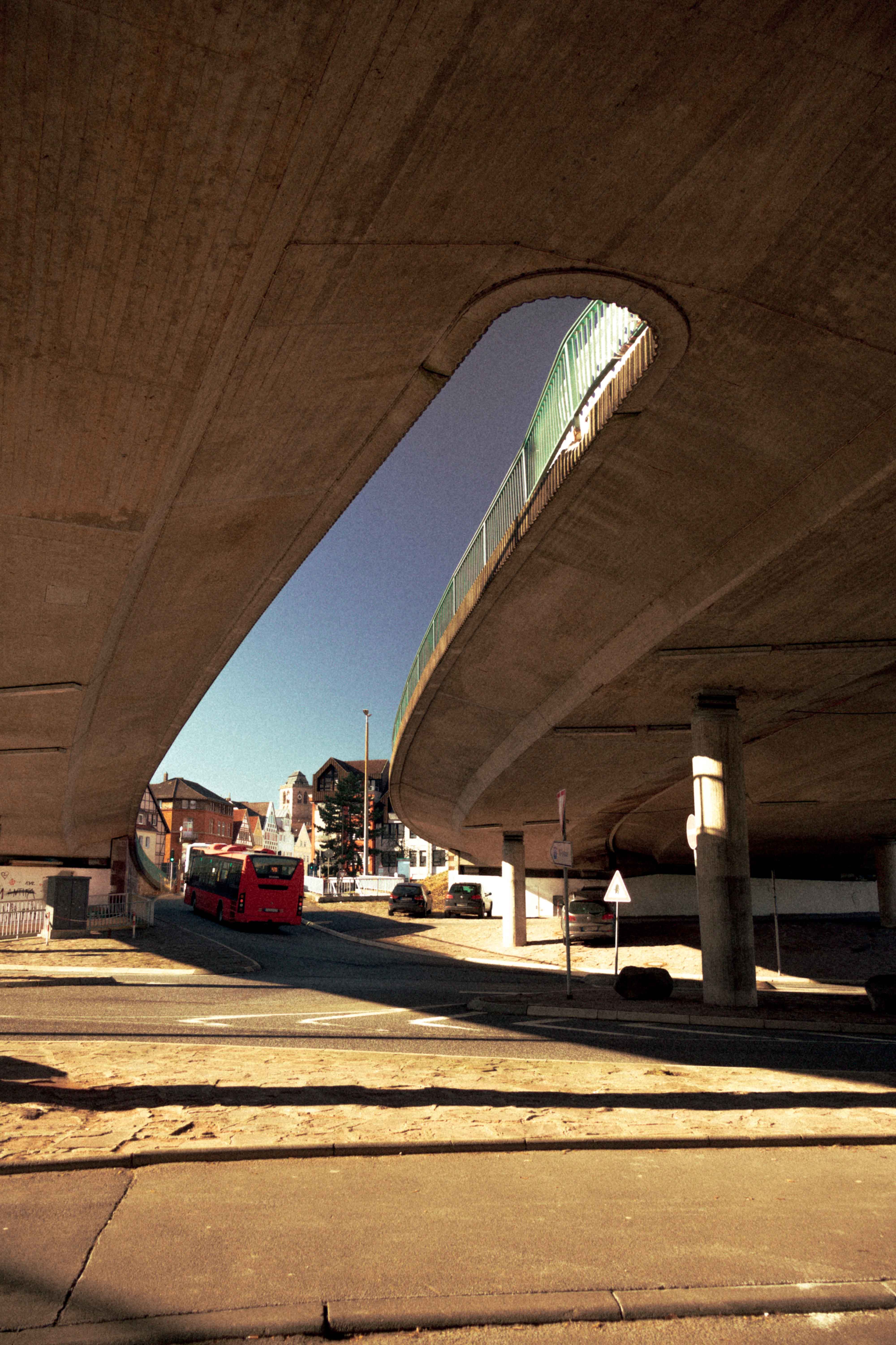 Urban scene with a red bus under a large concrete bridge on a clear day.