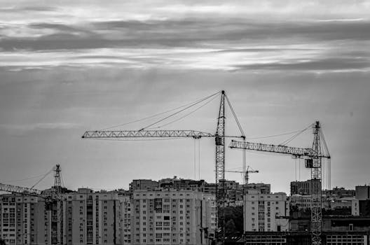 Black and white photo of tower cranes at city construction site against cloudy sky.