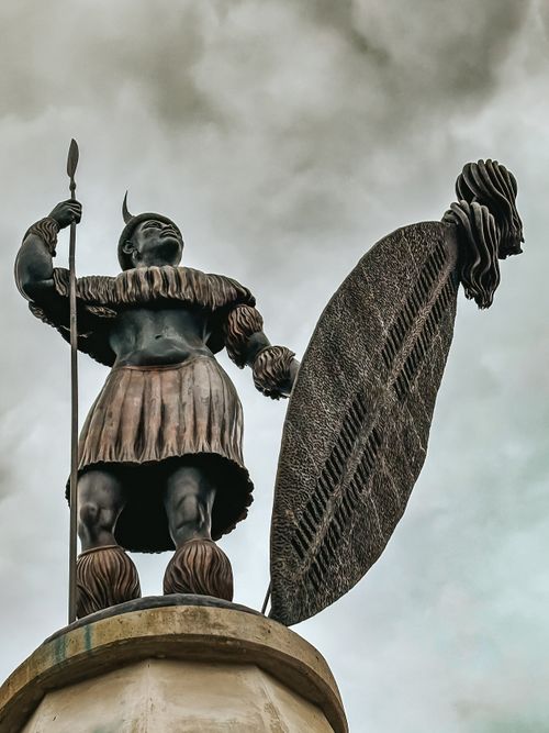 Free Impressive statue of a Zulu warrior with spear and shield under a cloudy sky. Stock Photo