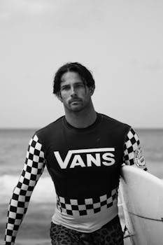 A black and white photo of a male surfer holding a surfboard by the ocean.