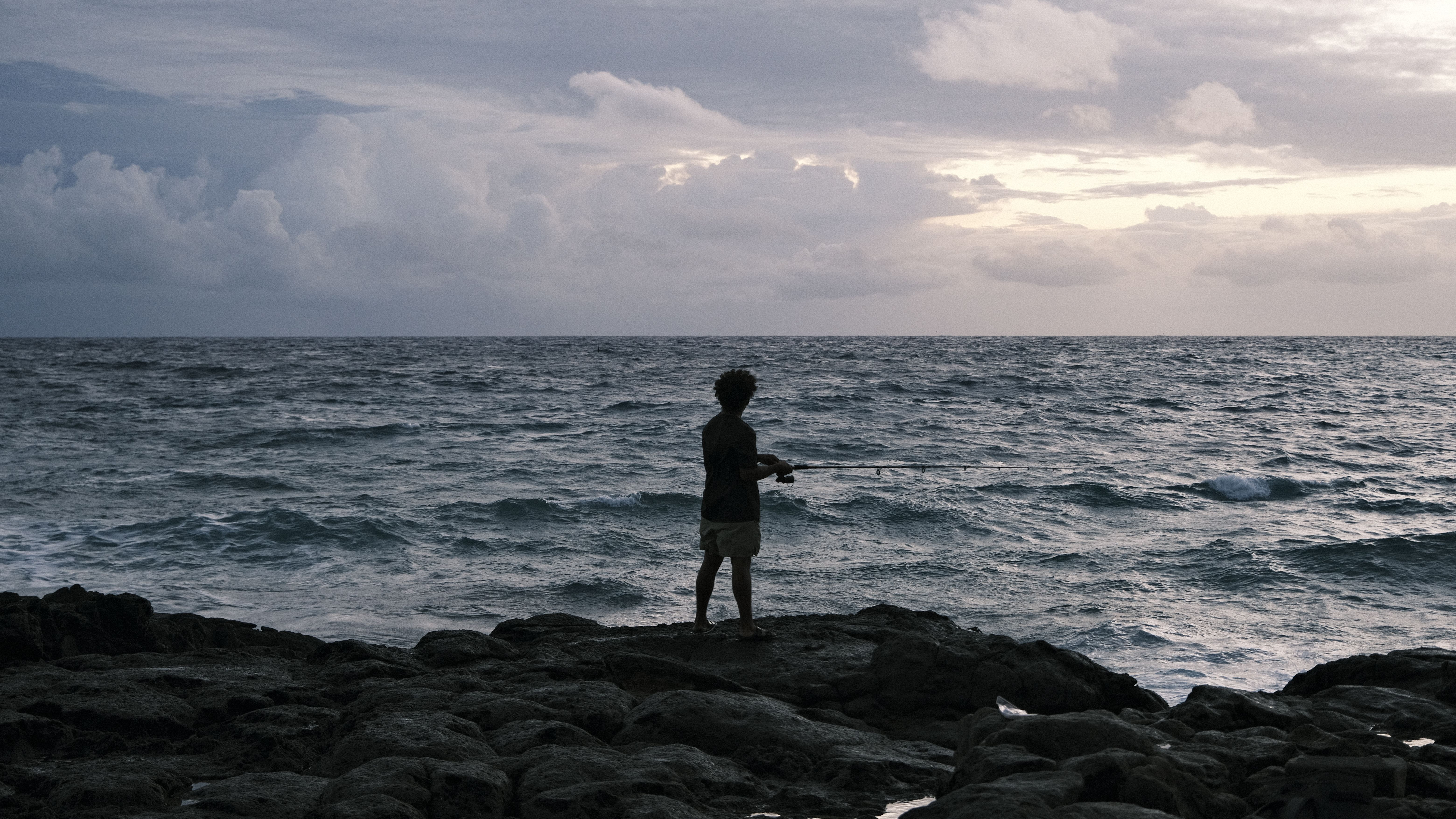 A man fishes alone on a rocky ocean shore under cloudy skies, at dusk.