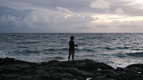 Free A man fishes alone on a rocky ocean shore under cloudy skies, at dusk. Stock Photo