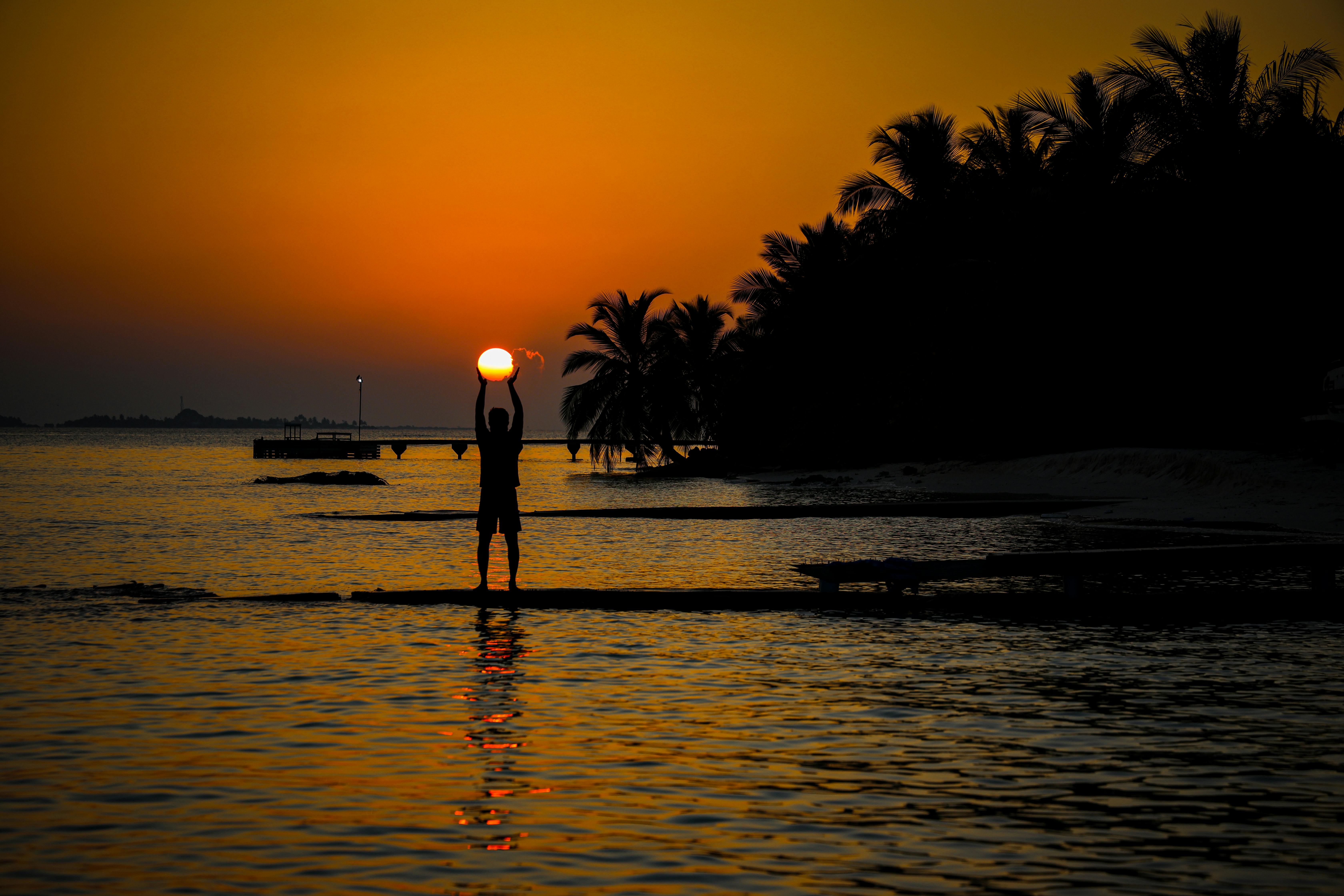 A serene tropical beach at sunset with a silhouetted person holding the sun, creating a magical scene.