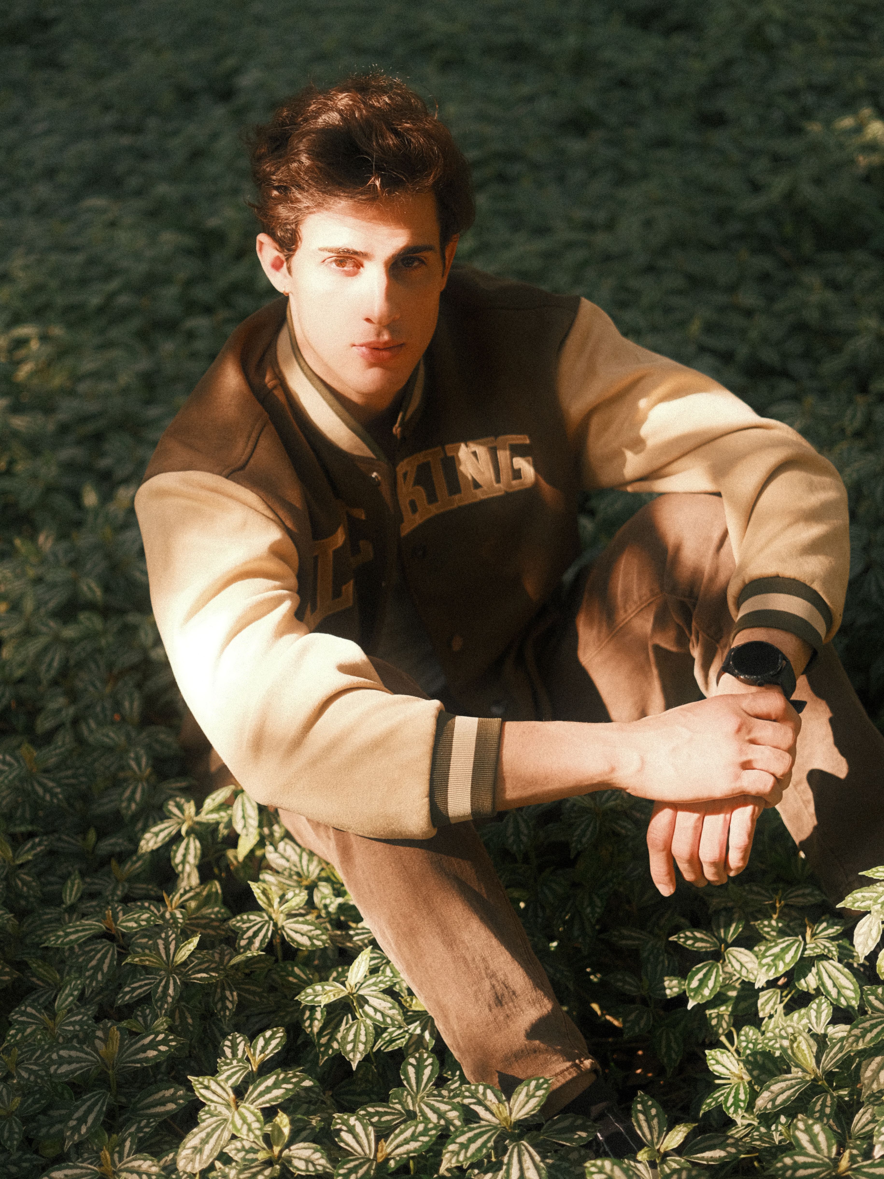 Young man sitting among green foliage, wearing a varsity jacket in natural light.