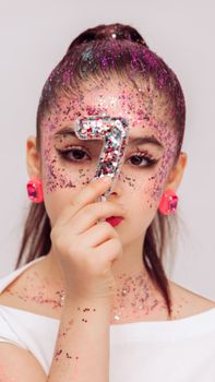 Close-up of a girl holding a number seven, covered in glitter, bright and artistic.