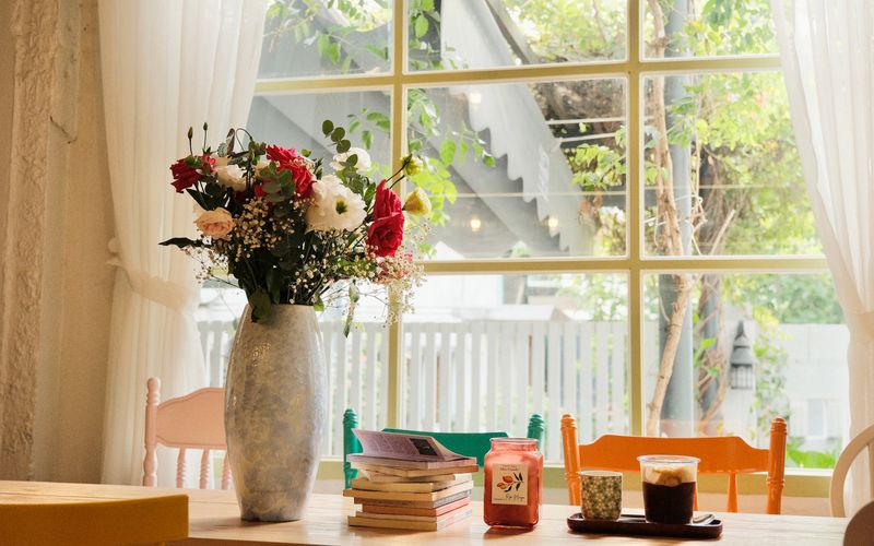 Cozy cafe table with flowers, books, and drinks by a bright window.