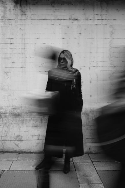 Black and white image of a woman standing against a wall with motion blur of passersby.
