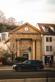 A historic building with a large clock, framed by trees, with a car passing by on a city street.