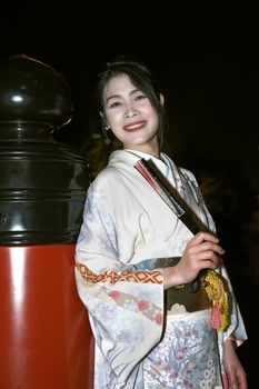 Elegant woman in a traditional kimono holding a fan, standing against a lantern at night.