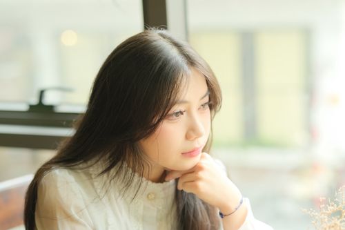Free Portrait of a young woman gazing thoughtfully out a window in soft indoor light. Stock Photo