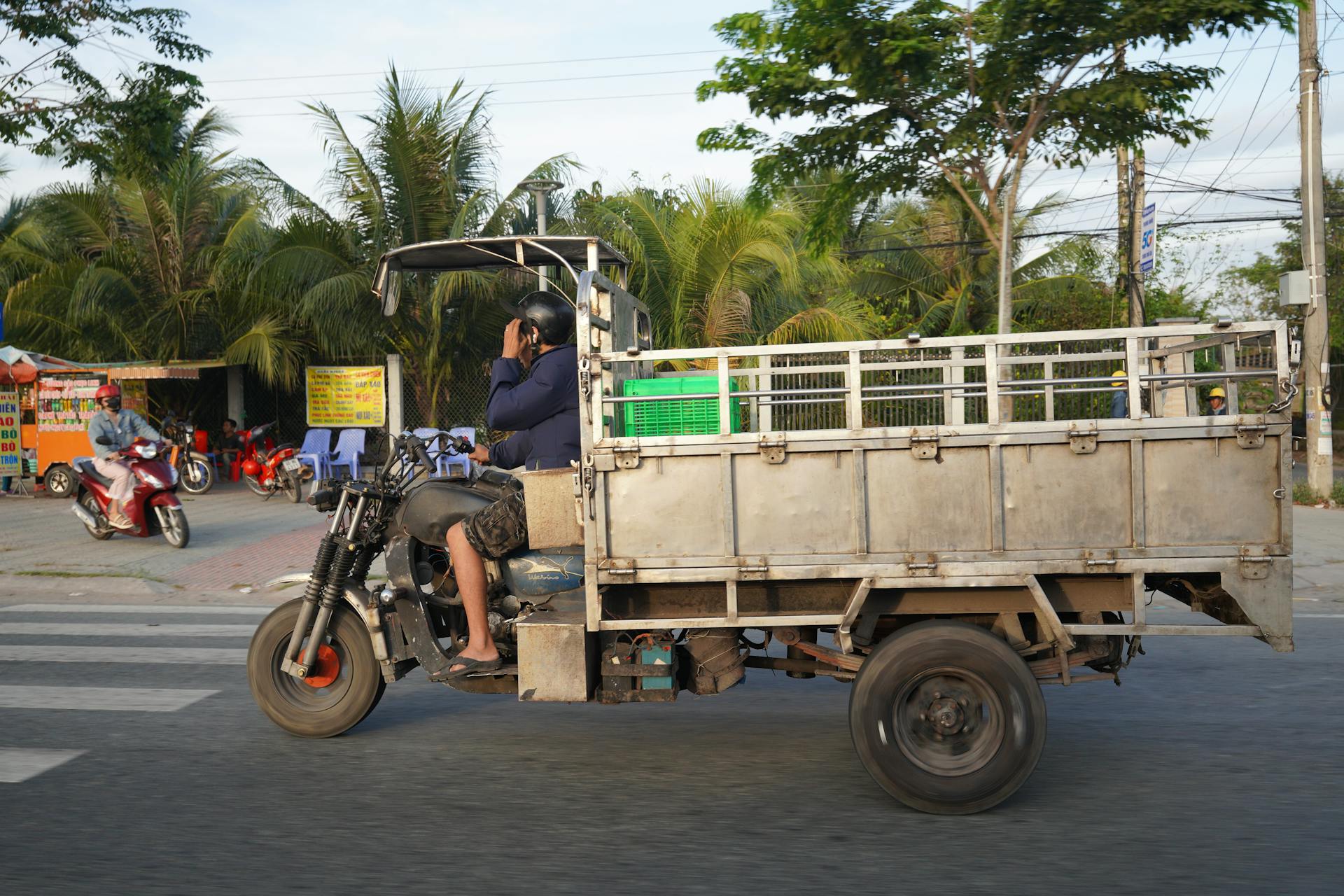 A man rides a customized motorcycle truck on a city street, surrounded by palm trees.