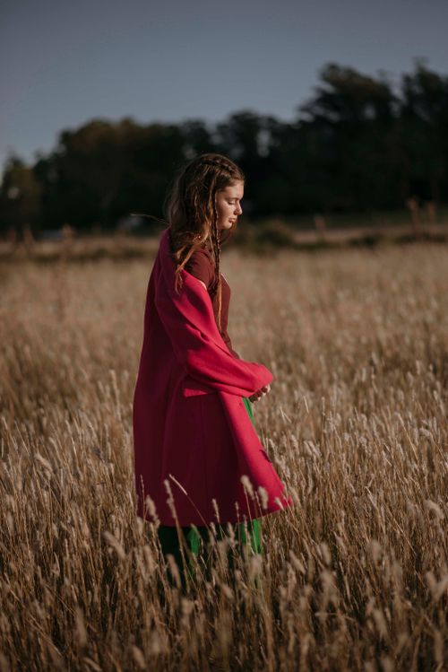 Free A contemplative woman in a red coat strolls through a serene autumn wheat field at sunset. Stock Photo