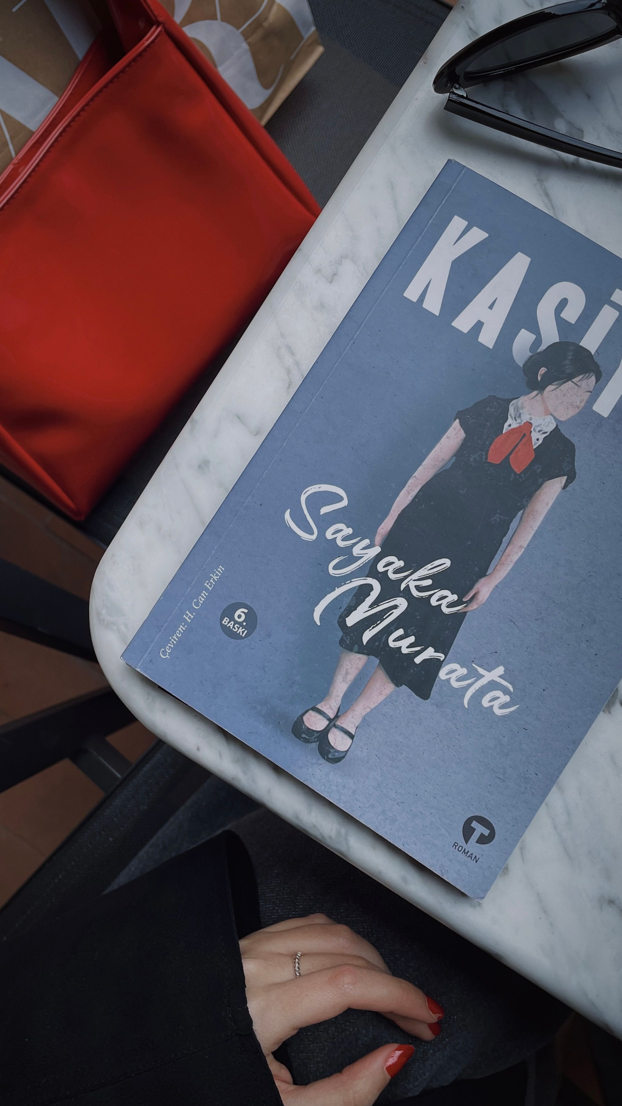 Flat lay of a book by Sayaka Murata with a red handbag and sunglasses on a marble table.