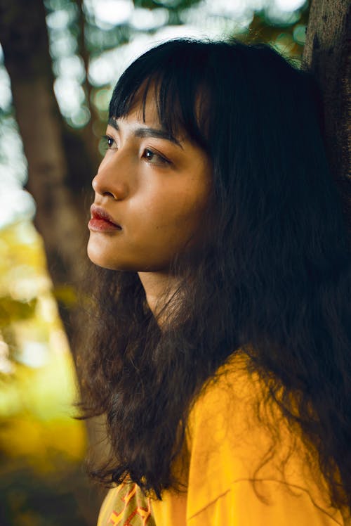 Side view of young pondering ethnic lady with makeup in bright wear looking away while standing near tree trunk