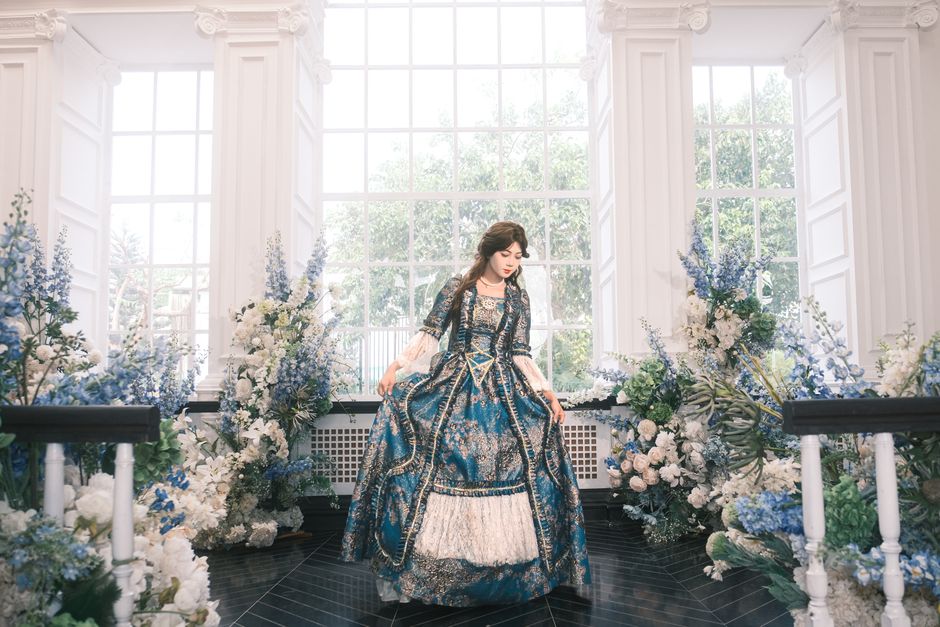 A woman in a blue Victorian dress stands amidst elegant floral decor indoors.