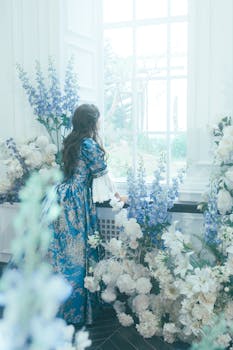 Elegant woman in blue floral dress gazes out a large window surrounded by flowers.