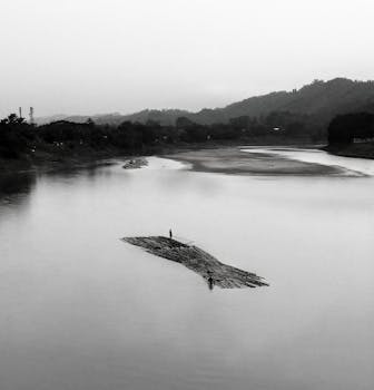 这是一张宁静的黑白图像，描绘了朦胧山丘背景下，一条宁静河流上的一叶孤筏