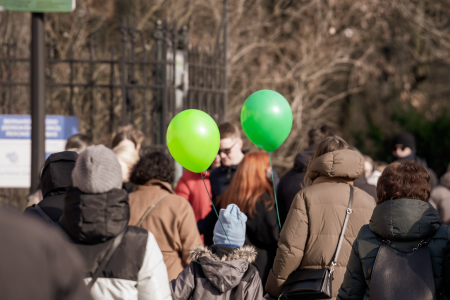 A crowd gathers in Vilnius park, holding bright green balloons during a sunny day.