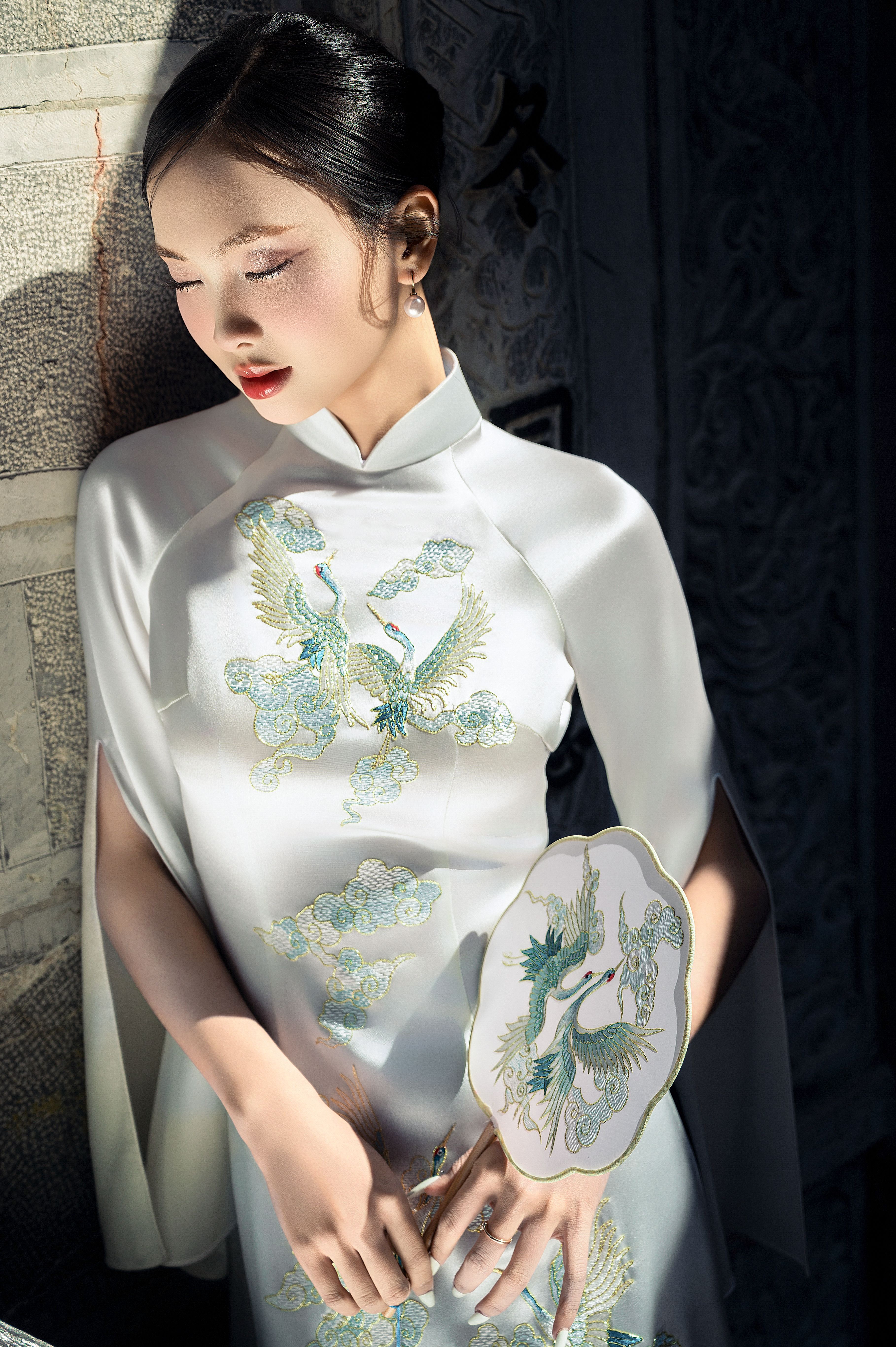 Woman in white dress with embroidery, holding decorative fan, in soft light.