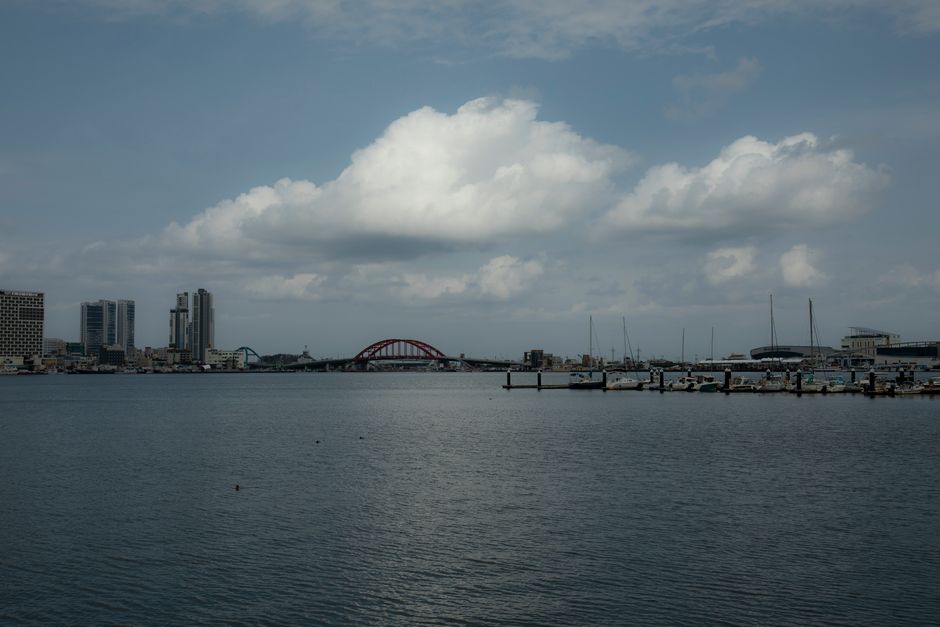 Serene view of a harbor with a distinctive red bridge and city skyline under a cloudy sky.