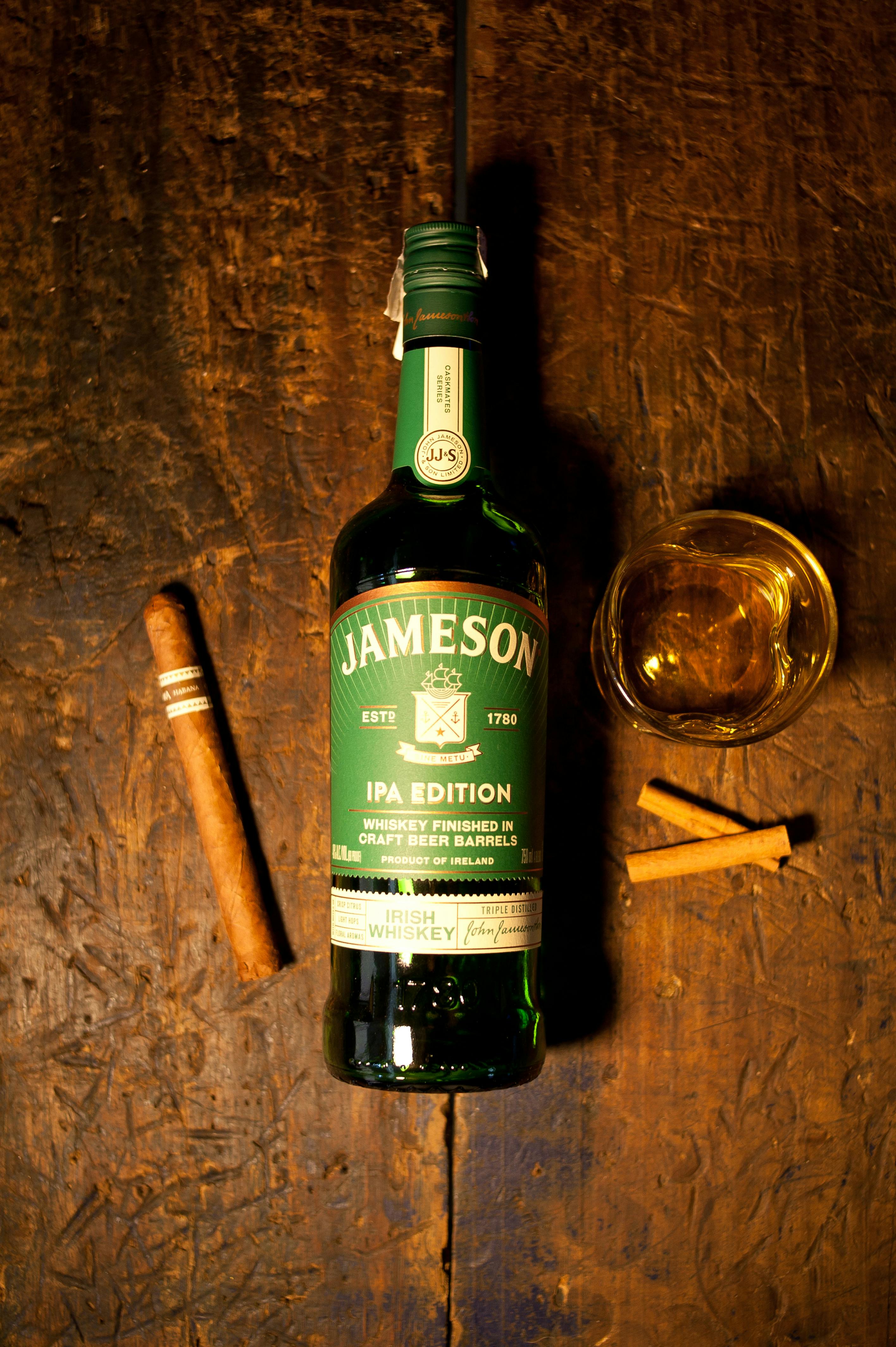 Jameson IPA Edition whiskey bottle, cigars, and glass on rustic wooden table.