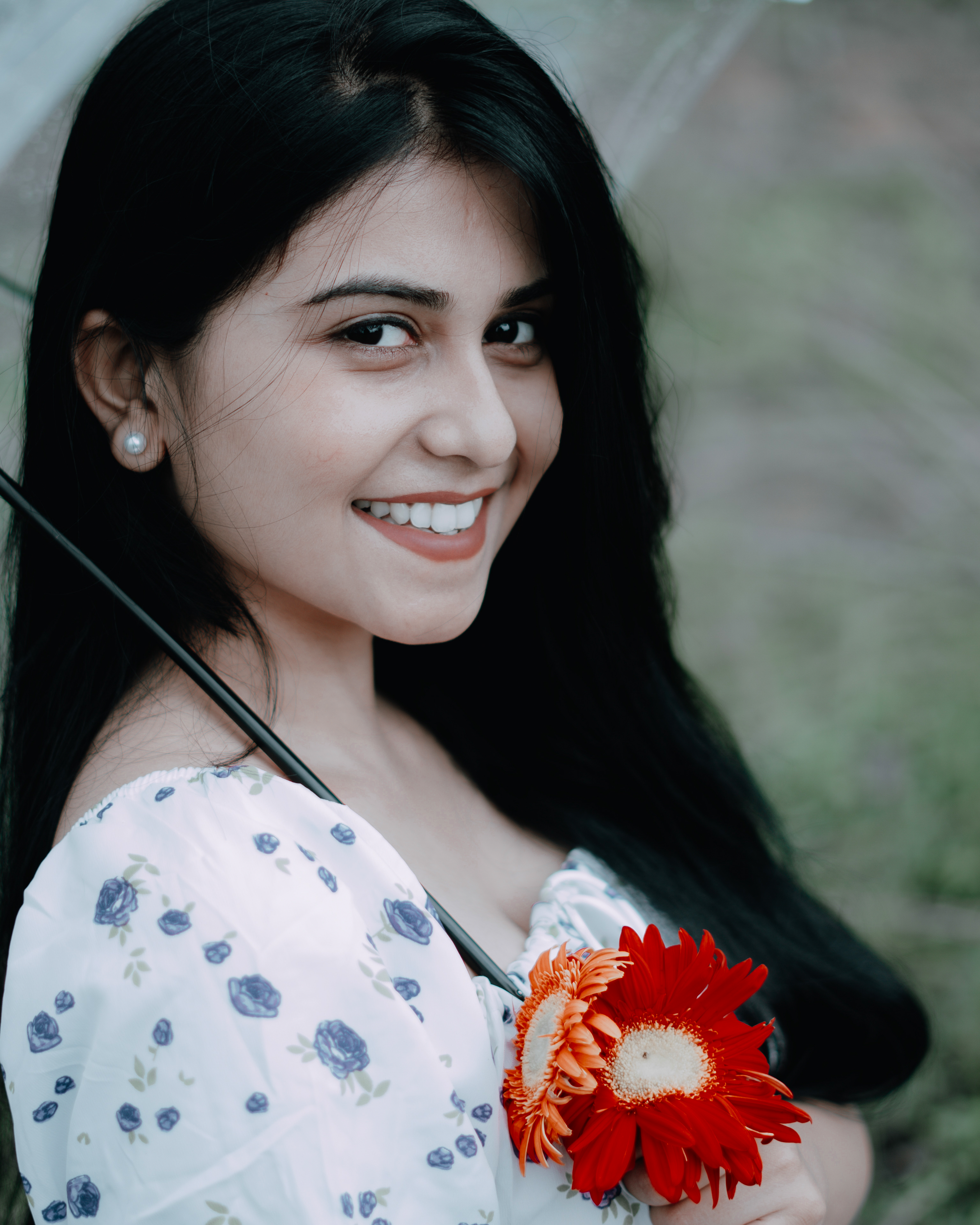 A cheerful woman in a floral dress holding a red gerbera flower and umbrella outdoors.