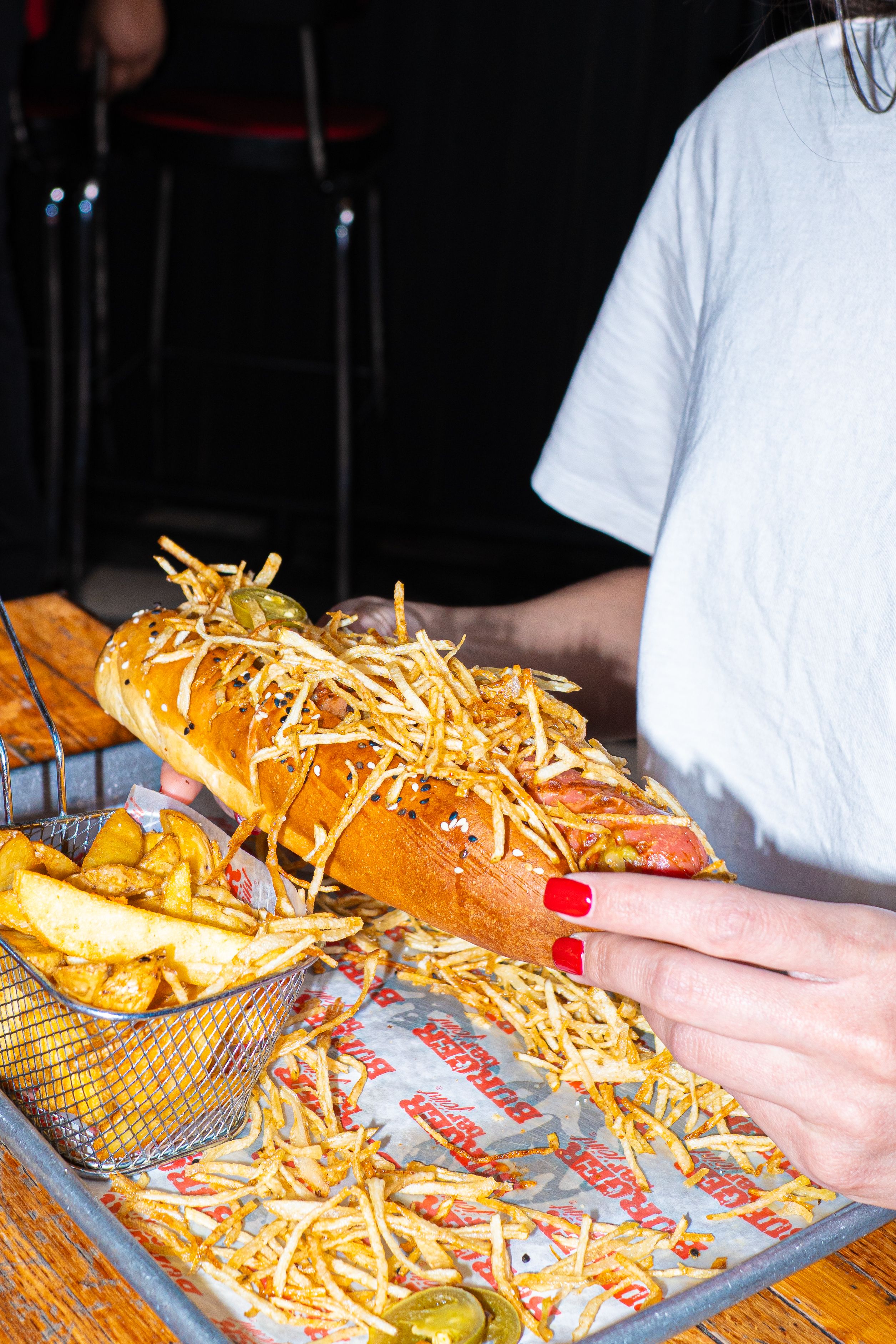 A close-up of a gourmet hotdog topped with fried potatoes, served with fries in a casual dining setting.
