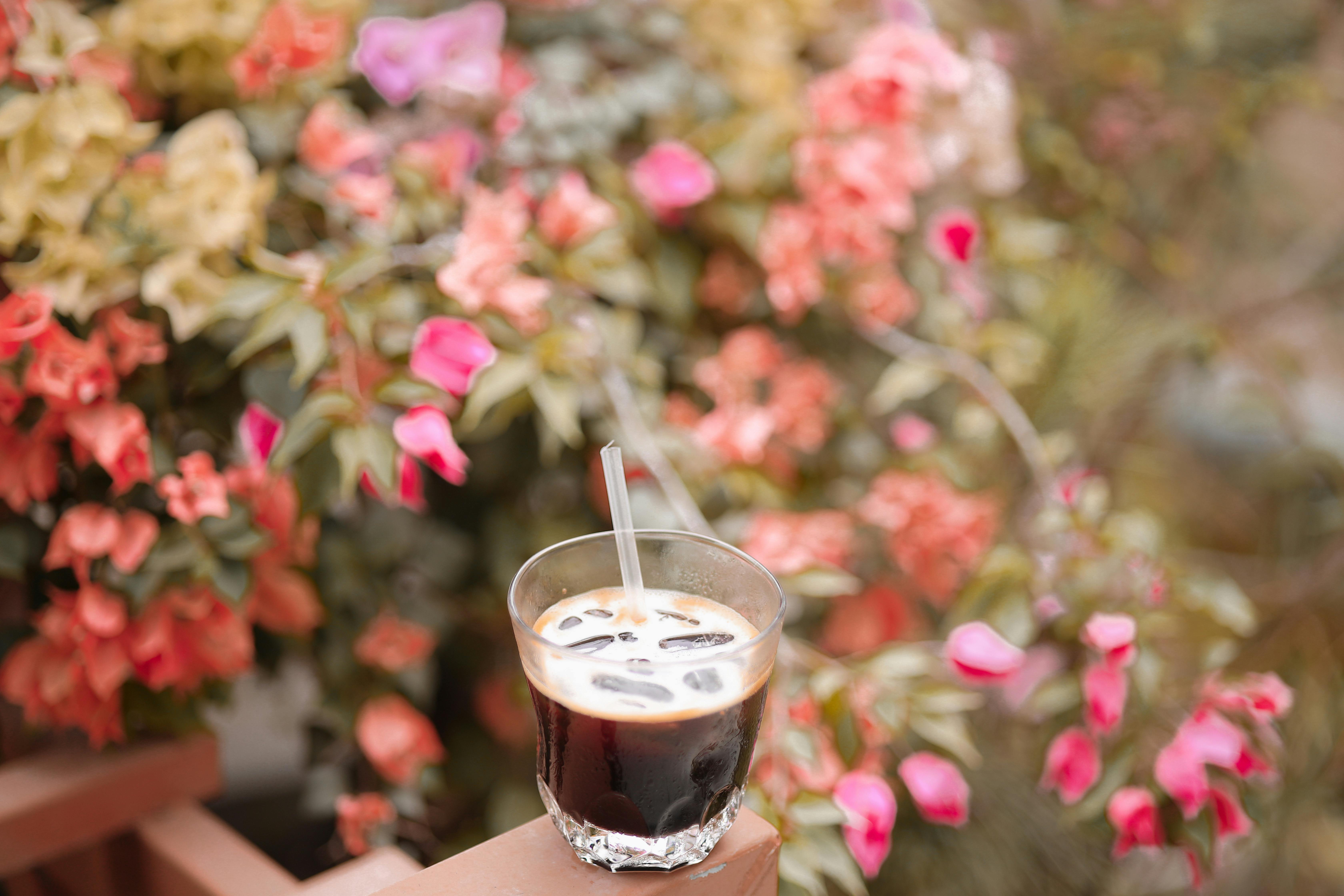 A glass of iced coffee on a railing set against colorful blooming flowers.