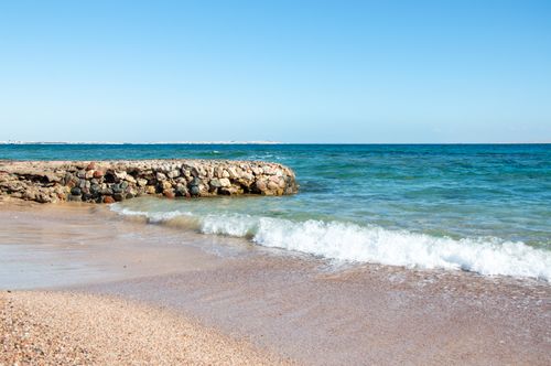 Free Peaceful beach scene with waves crashing against a stone breakwater under a sunny blue sky. Stock Photo