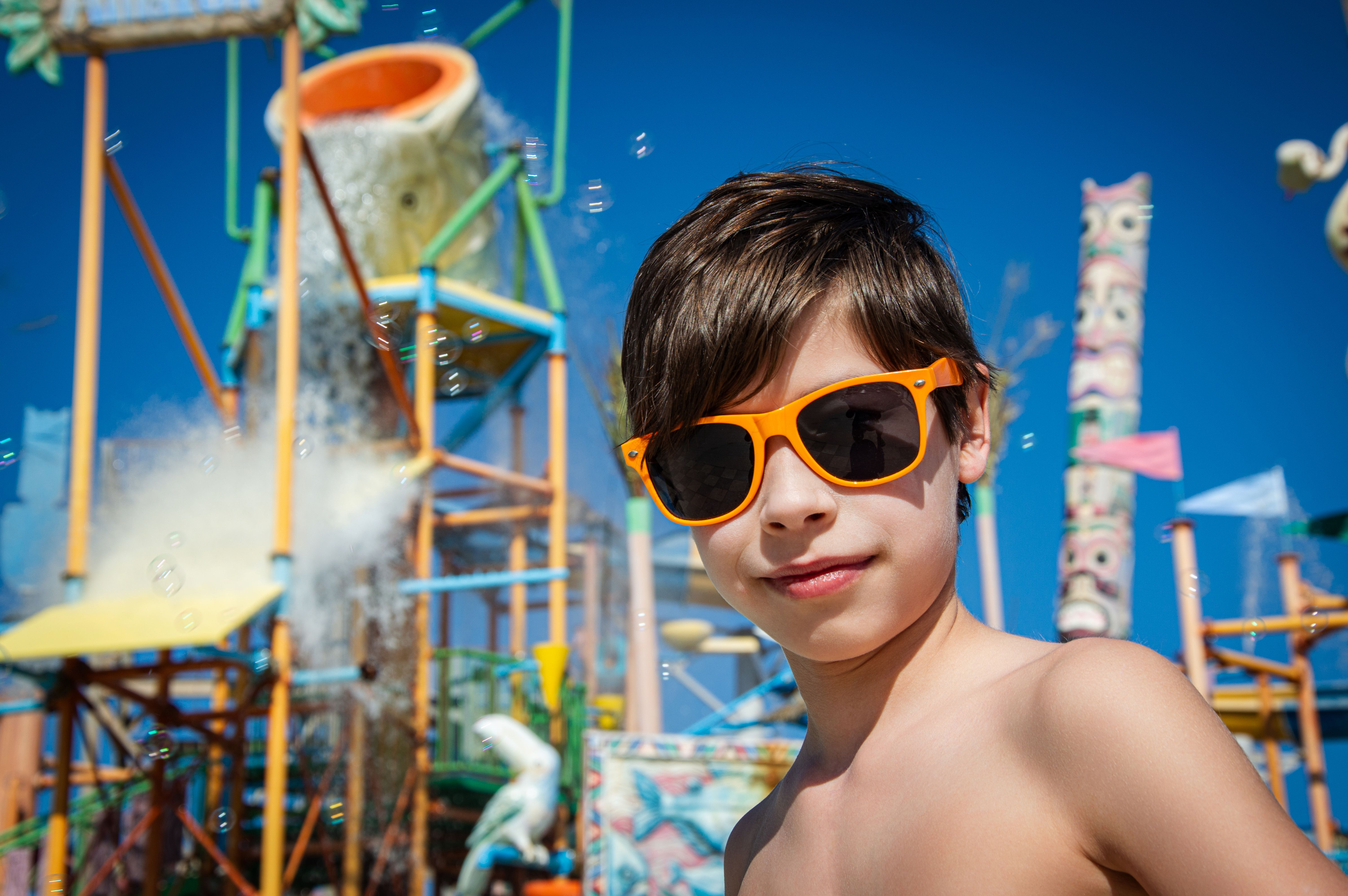 A young boy wearing sunglasses enjoys a sunny day at a colorful water park.