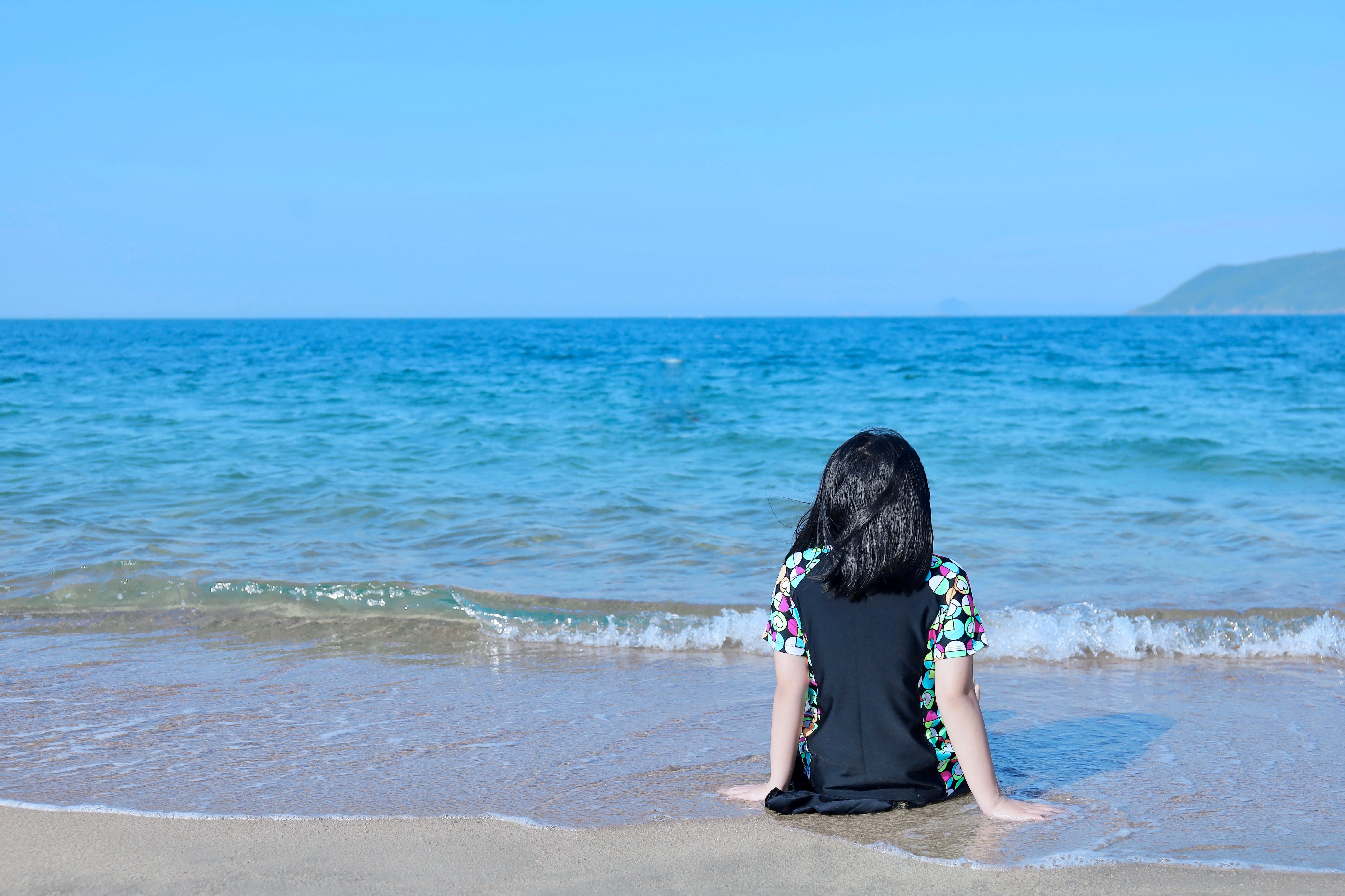 Young girl sitting on beach facing the ocean, enjoying a sunny day.