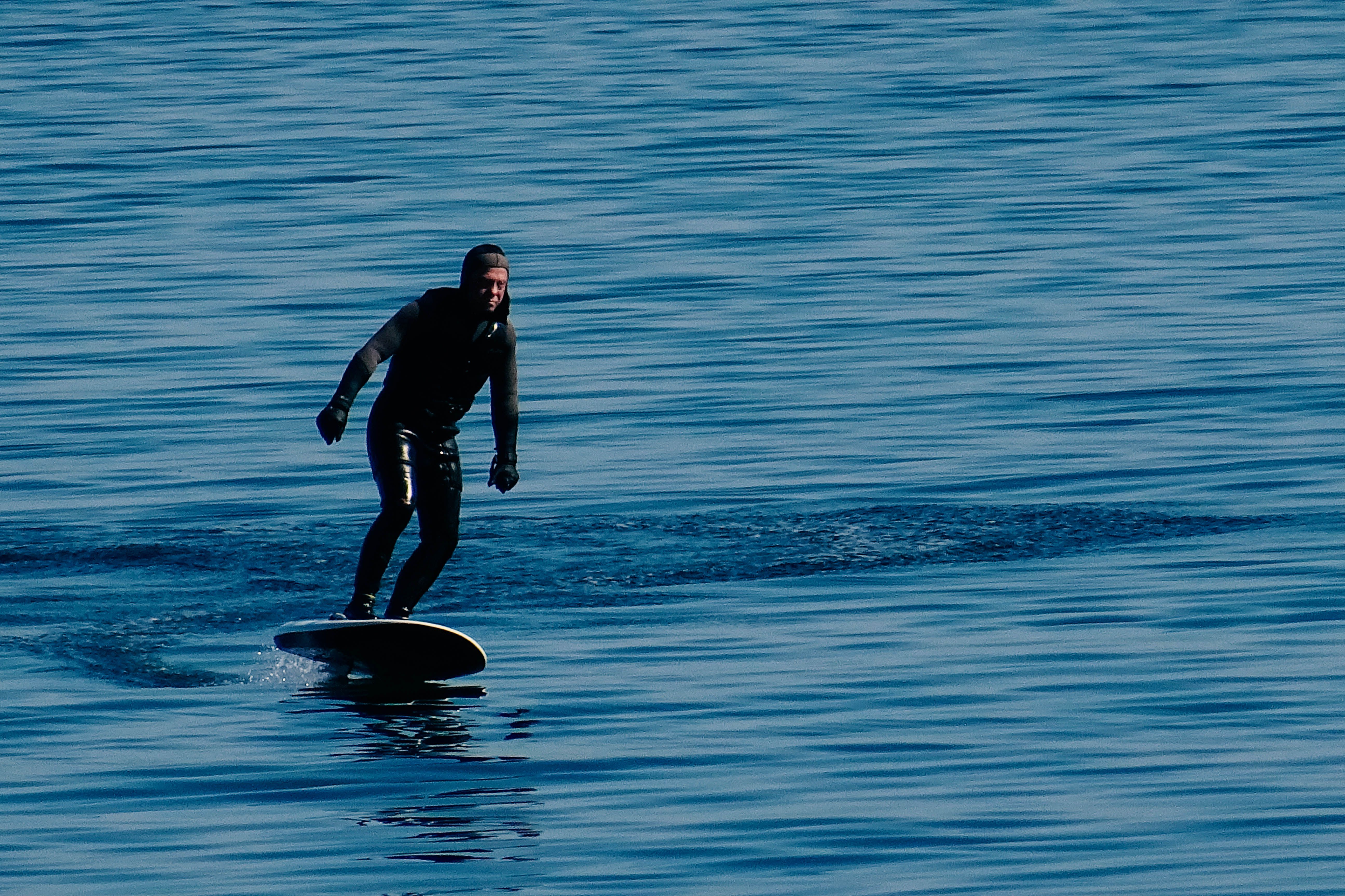 A man in a wetsuit surfs on a hydrofoil board across calm blue waters during the day.