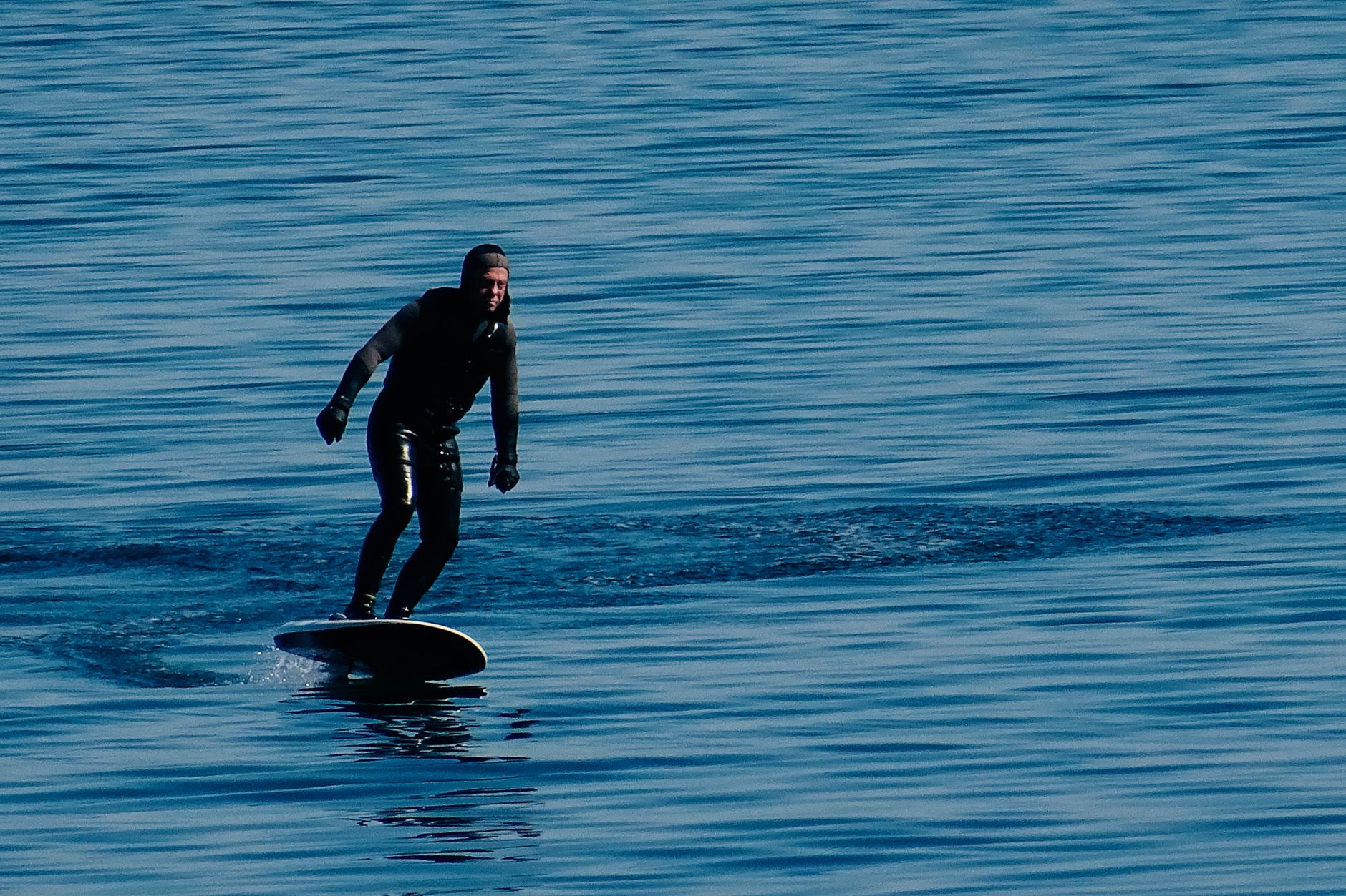 A man in a wetsuit surfs on a hydrofoil board across calm blue waters during the day.