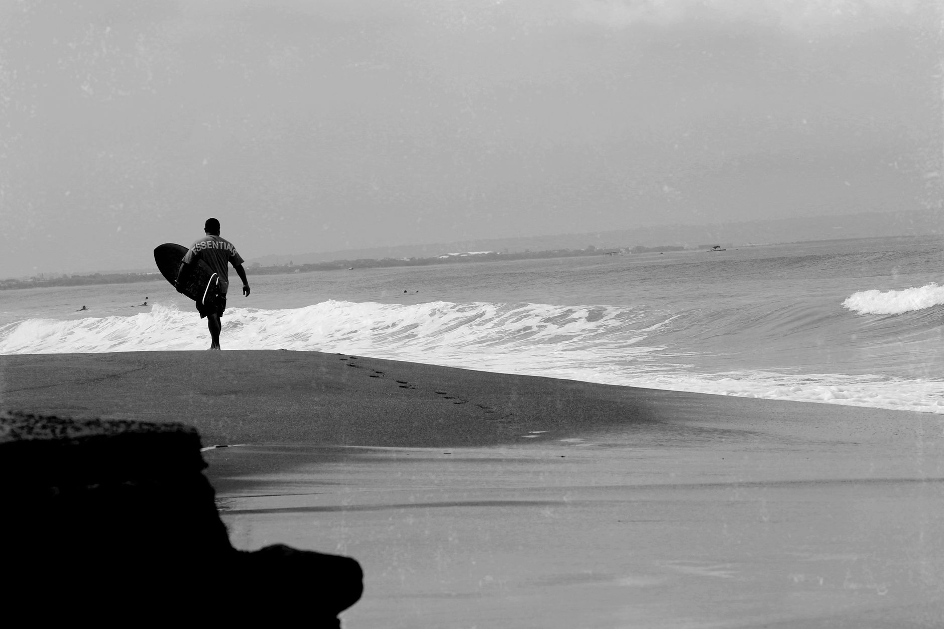 Black and white photo of a surfer walking on a Bali beach with a surfboard.