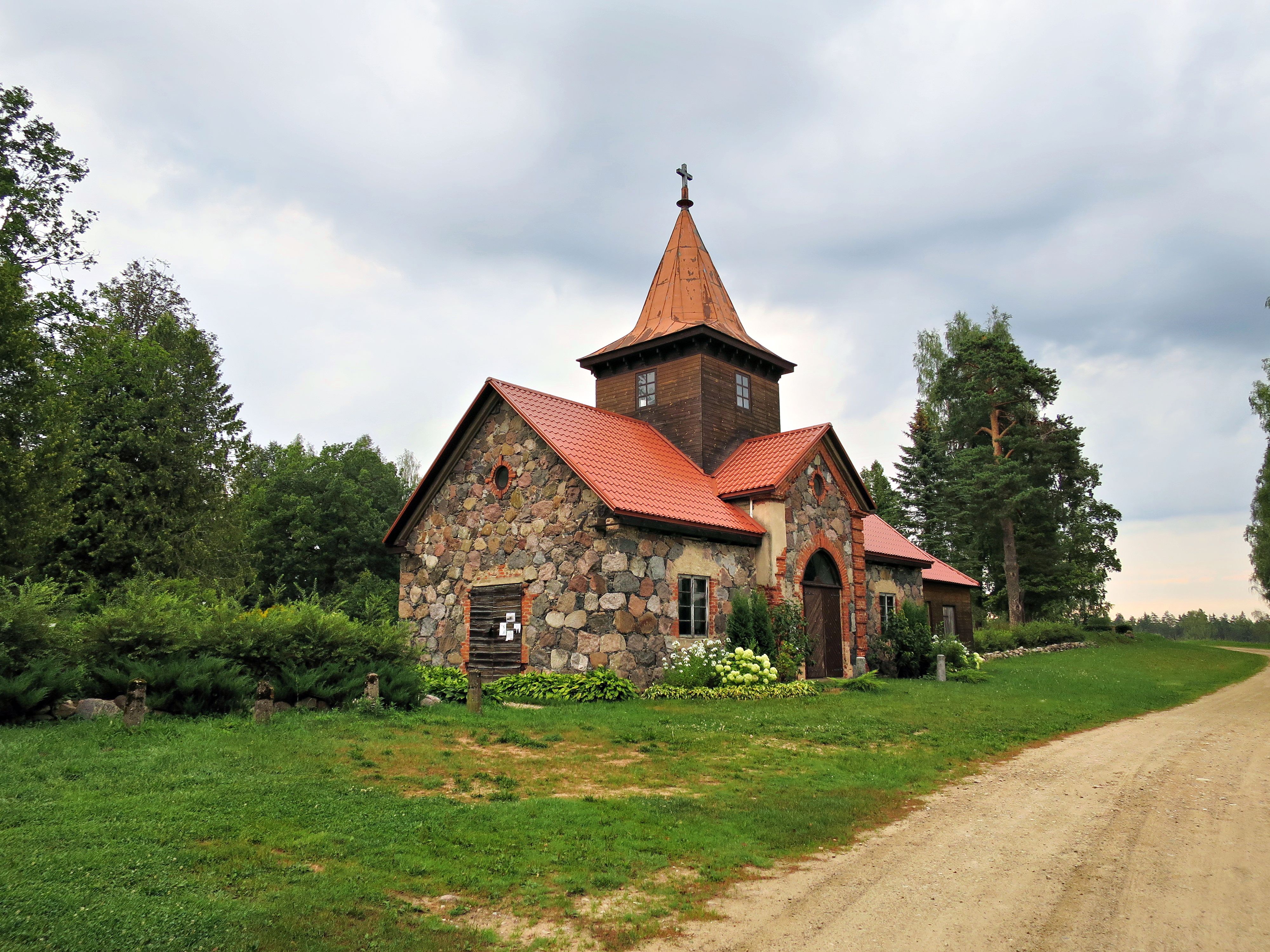 Quaint stone chapel with red roof in the serene Latvian countryside under cloudy skies.