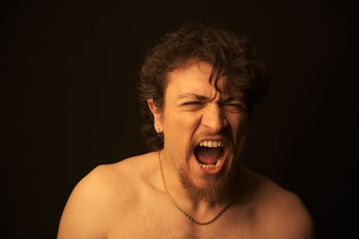 A bare-chested man with curly hair shouting passionately against a dark background.