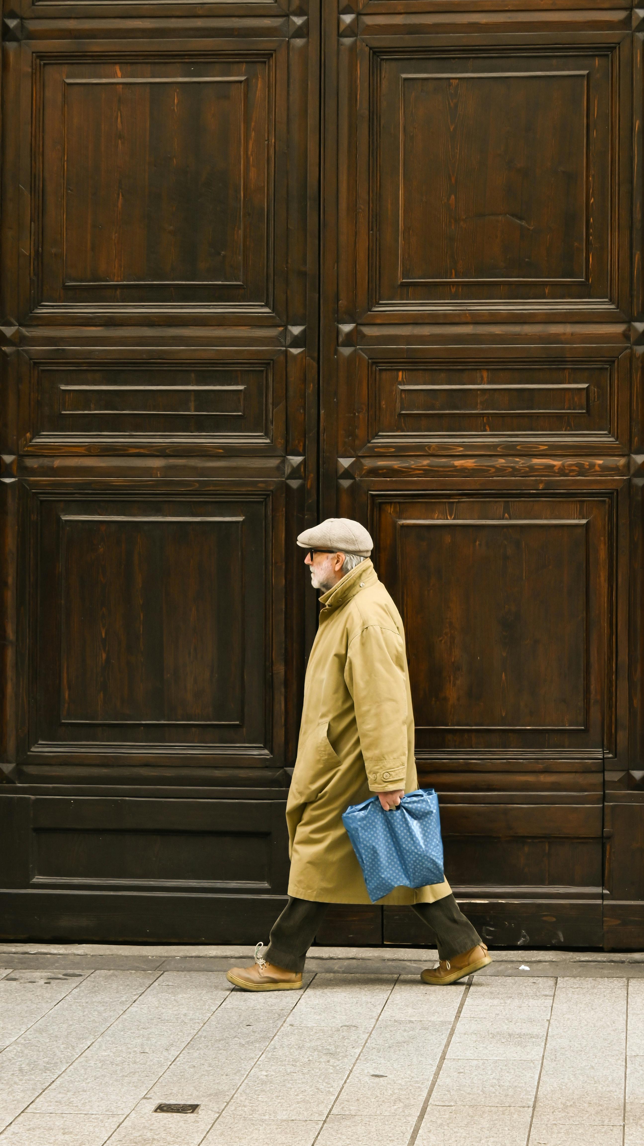 Elderly man in a coat walks by a large wooden door carrying a blue bag.