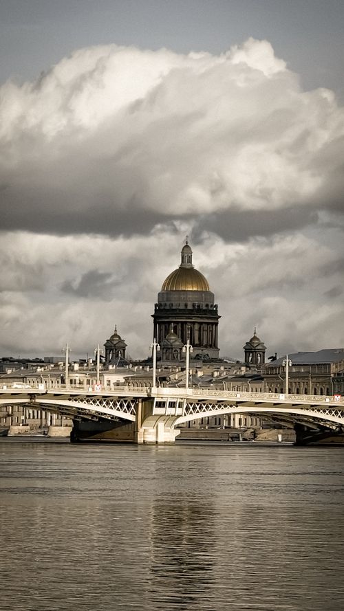 Free Majestic view of St. Isaac's Cathedral under dramatic clouds with a serene bridge spanning the river. Stock Photo