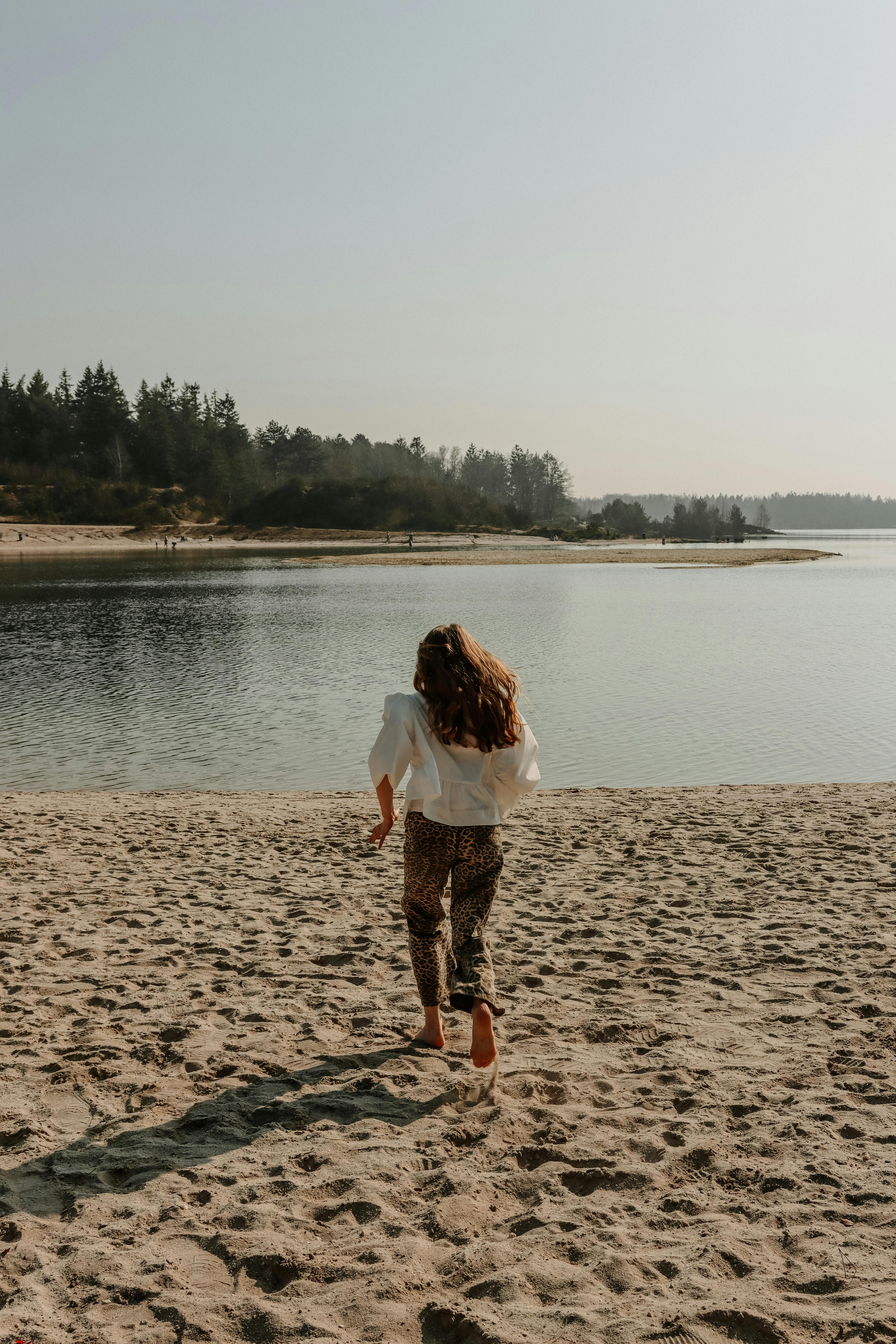 A woman runs towards a tranquil lake on a sandy beach in Drouwen, Netherlands, under a clear sky.