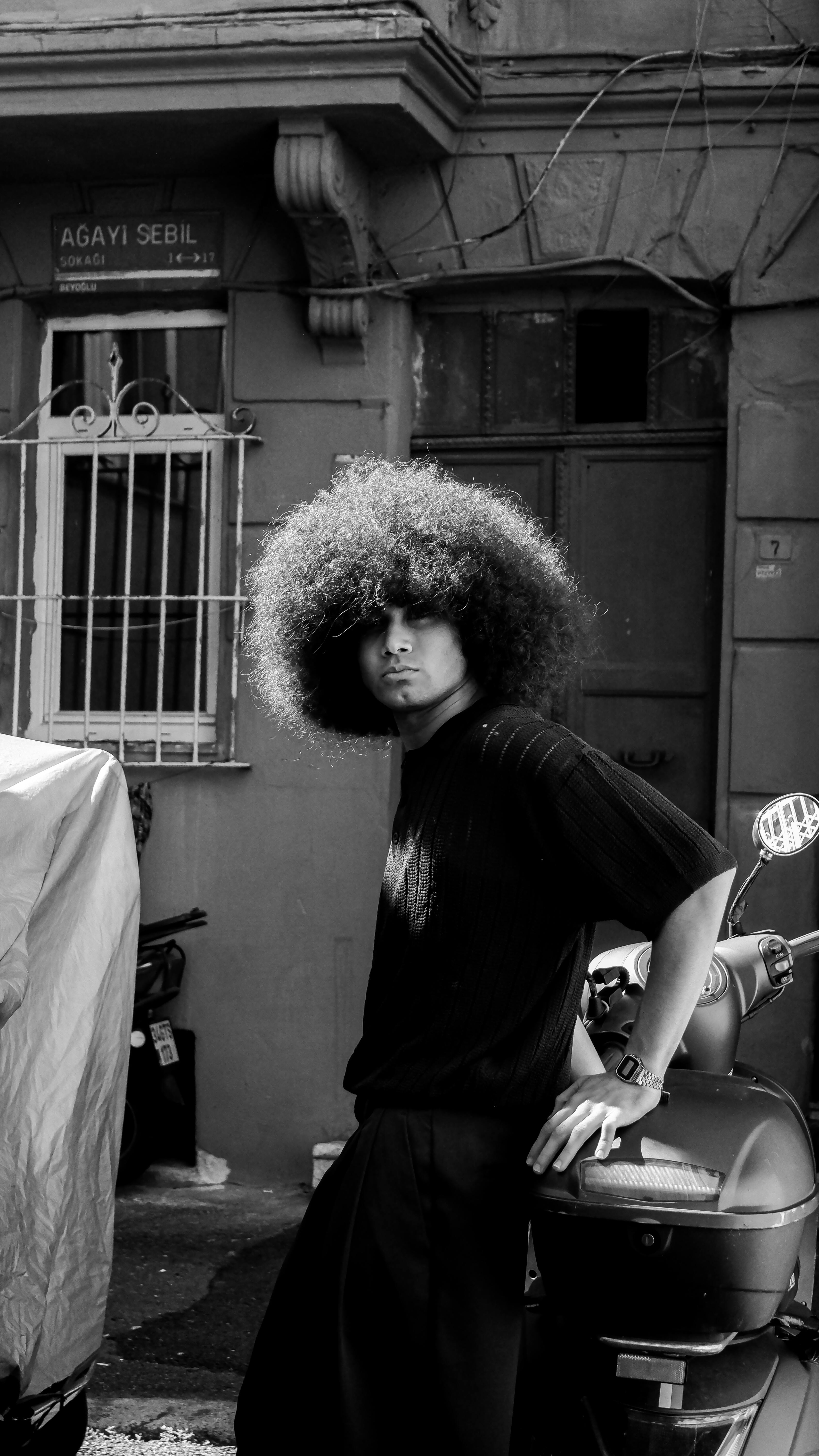 Black and white portrait of a man with afro hairstyle leaning on a scooter outdoors.
