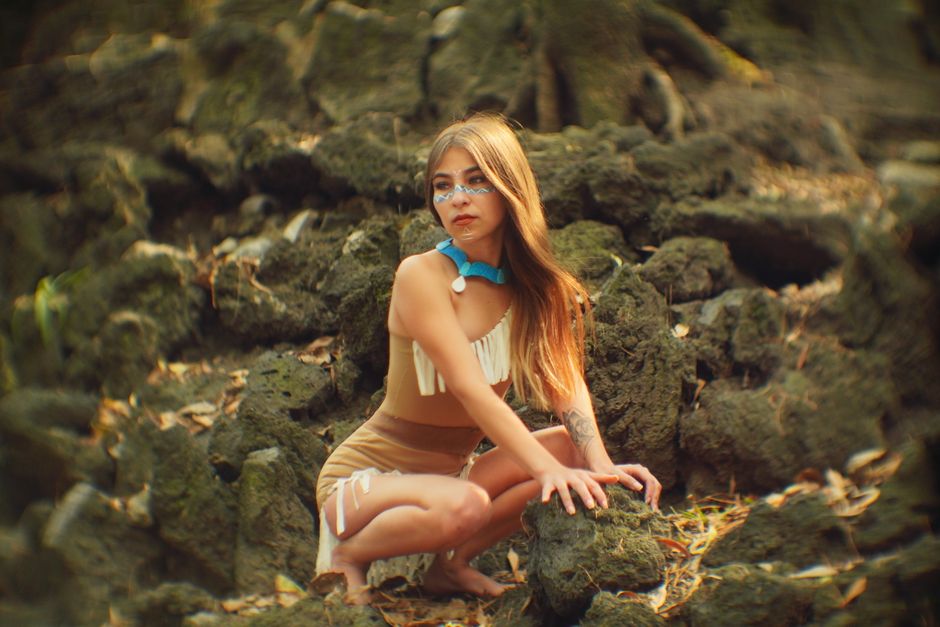A young woman in tribal costume poses amidst nature with colorful makeup.