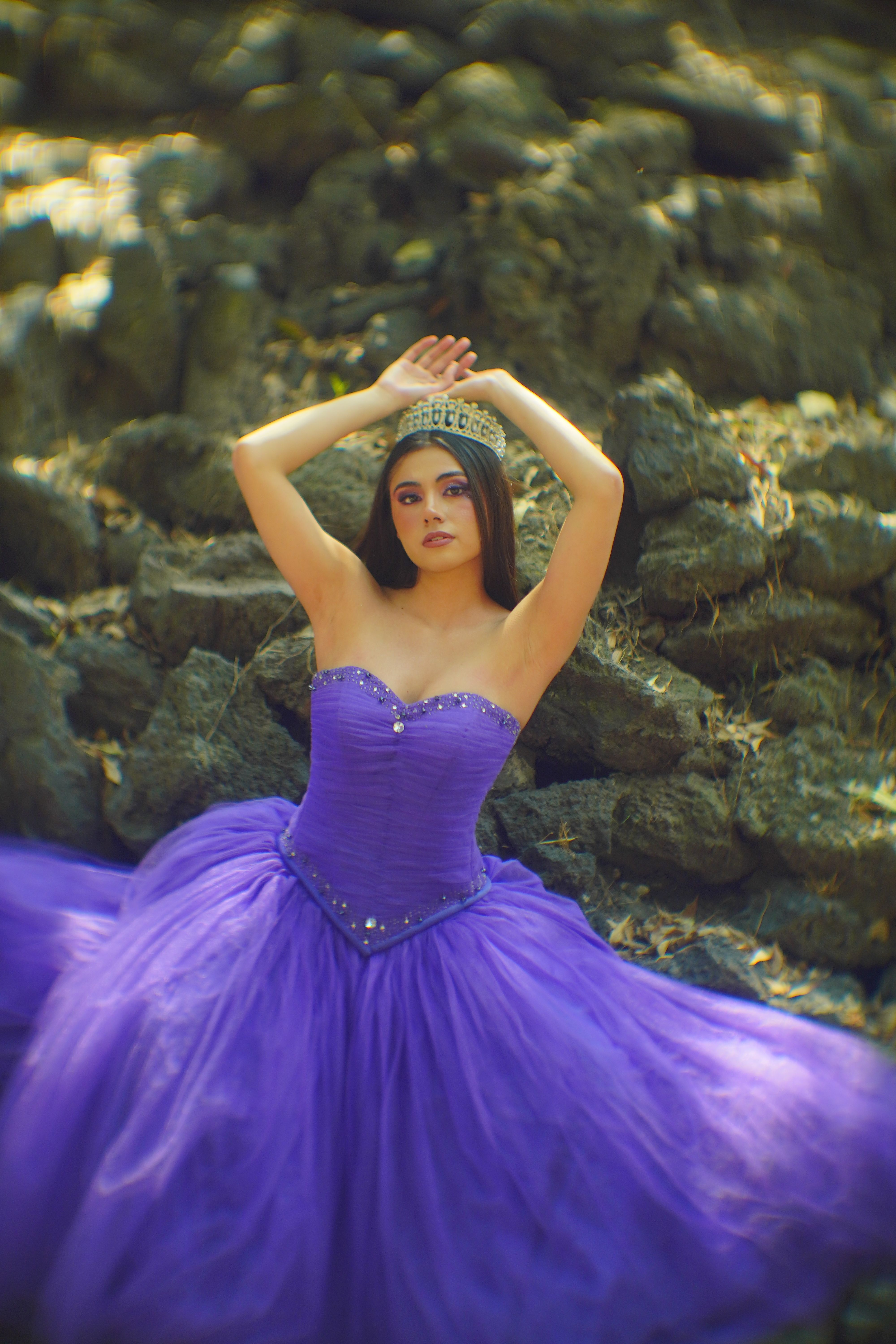 A woman in a purple gown and tiara poses elegantly outdoors on rocky terrain.