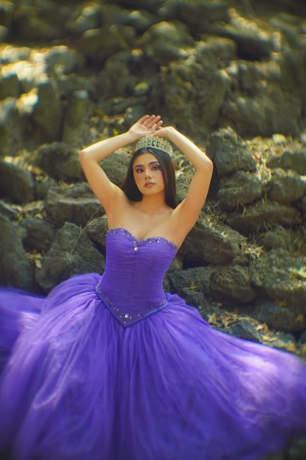 A woman in a purple gown and tiara poses elegantly outdoors on rocky terrain.