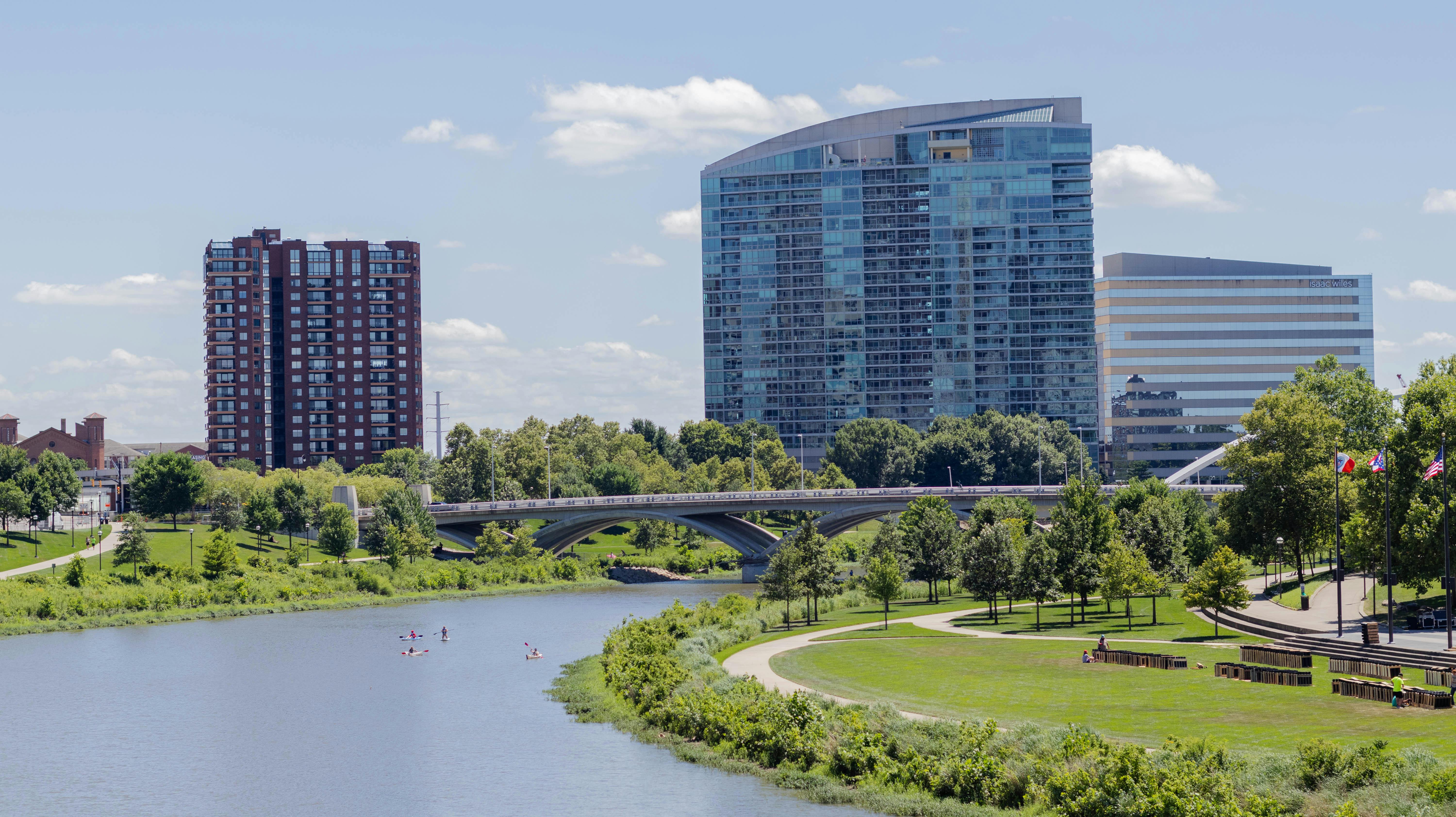 Scenic view of a modern urban skyline by a riverside with lush greenery and blue sky.