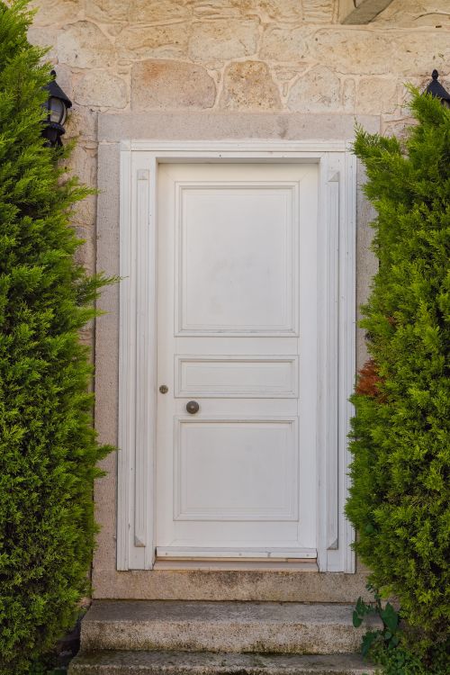 Free A classic white door framed by lush green bushes, offering a serene outdoor setting. Stock Photo