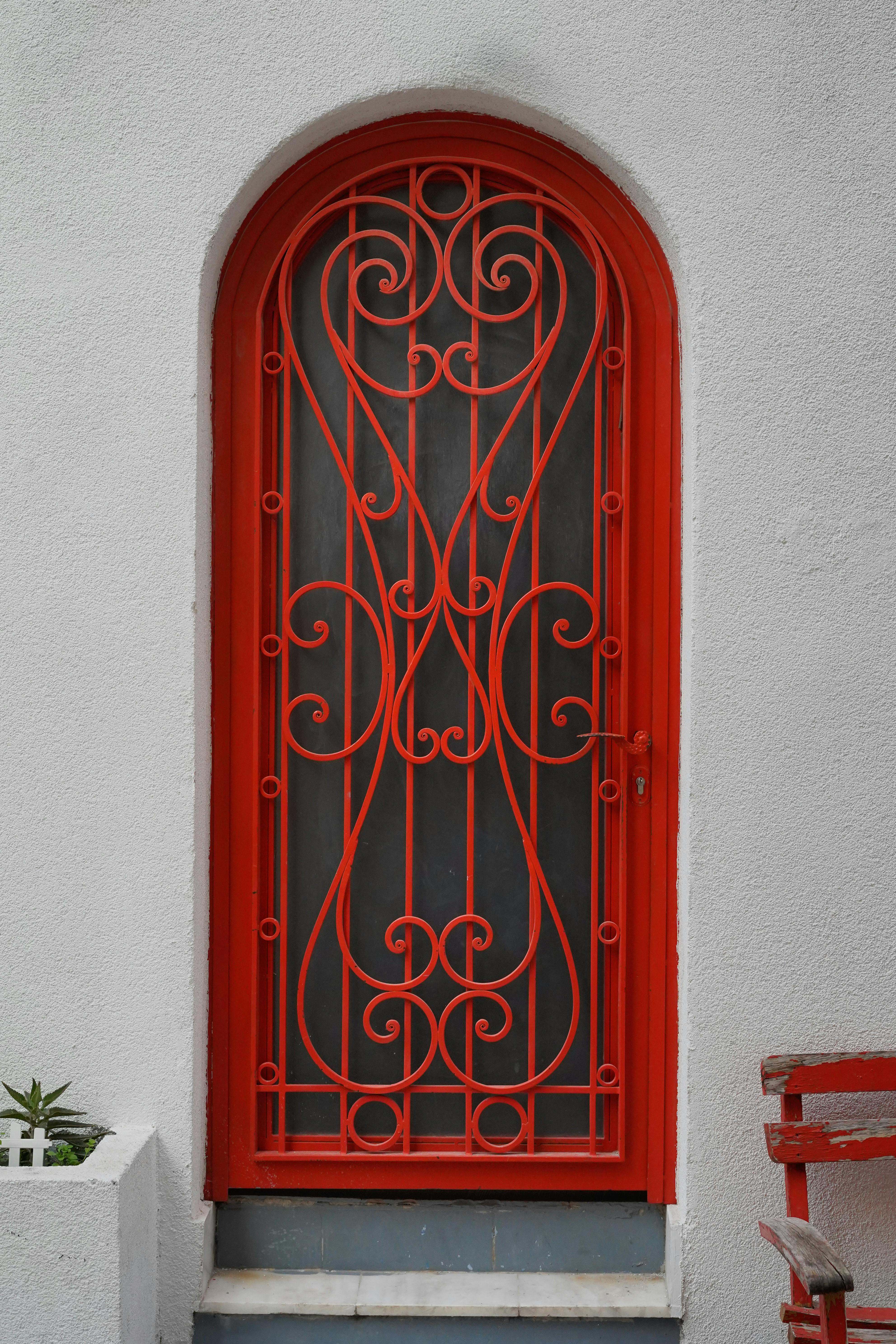 A striking red wrought iron door with intricate design on a white textured wall.