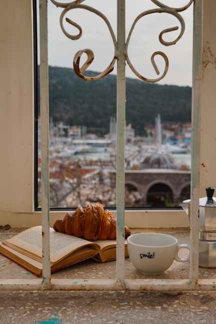 Croissant, coffee, and book by a window with a Mediterranean view.