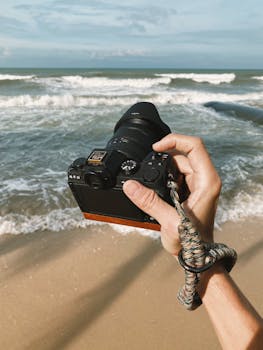 Hand of photographer holding camera, capturing ocean waves on a sunny day at a sandy beach.